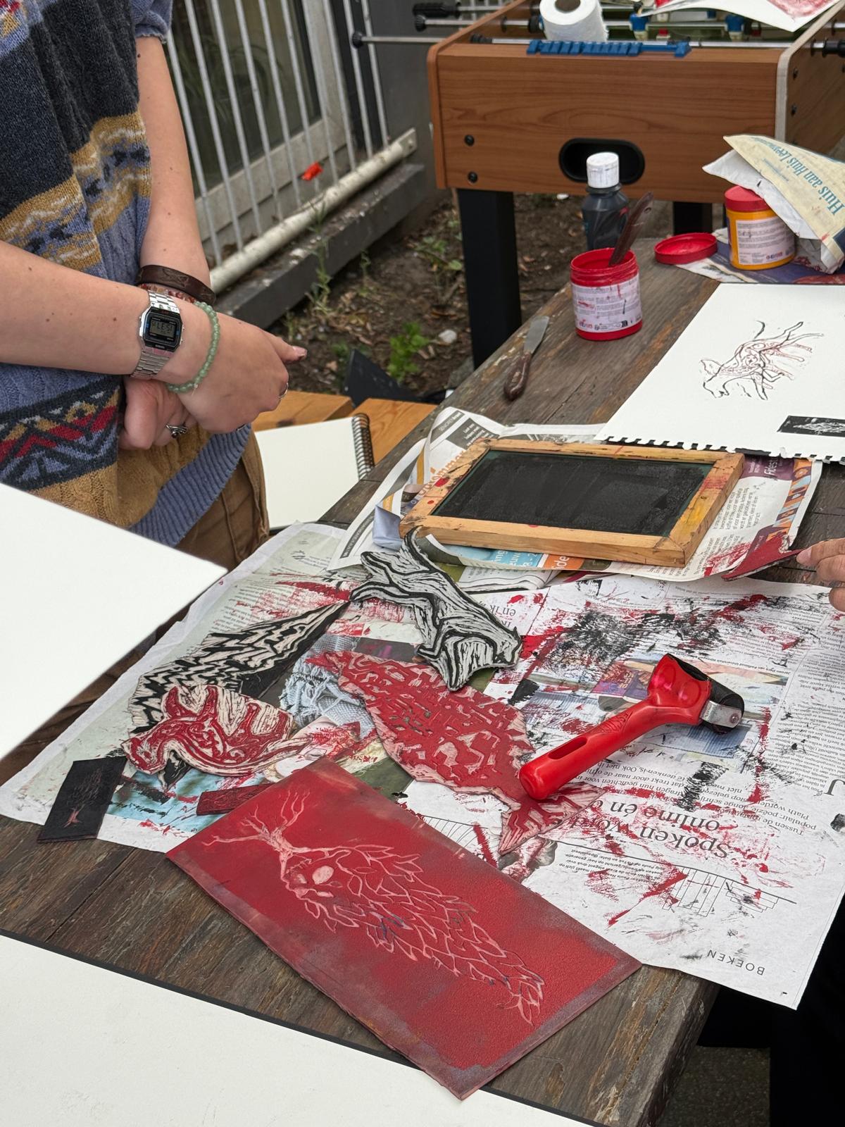 The kiosk table mid-session: carved lino blocks, red ink and a roller, freshly pulled prints laid out to dry.